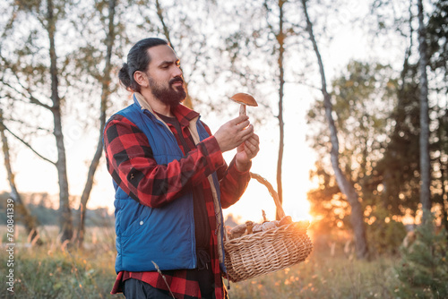 A mushroom picker man with a basket holds a mushroom in his hands in the forest. photo