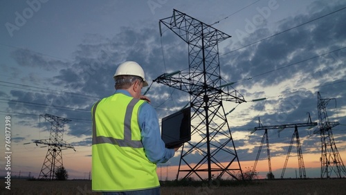 Engineer checks equipment with laptop walking to power transmission lines in sunset rural field backside view. Employee in helmet carries laptop at power generation plant in evening country meadow