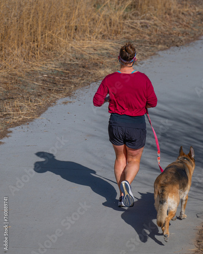 Woman wearing a mauve colored t-shirt with blue shorts jogging on a concrete paved trail  on a sunny autumn day with her German Shepard dog on a leash.