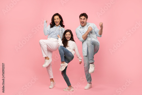 Overjoyed young family of three gesturing yes, celebrating success or win, man, woman and child girl shaking fists together on pink studio wall, full length