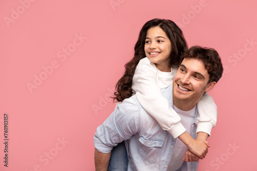Happy european young father carrying his daughter piggyback and looking at free space, posing over pink studio background, place for advertisement