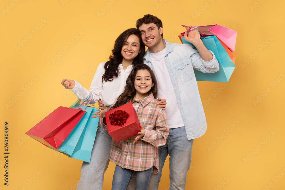 Cheerful family of three people holding bright shopper bags, excited teen girl holding wrapped present box, posing isolated on yellow studio wall Cheerful family of three people holding bright shopper bags, excited teen girl holding wrapped present box, posing isolated on yellow studio wall