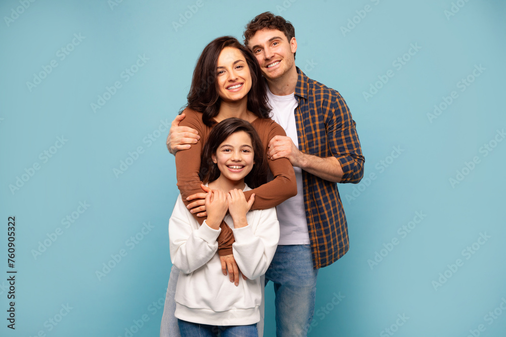 Healthy family relationship. Portrait of young dad and mommy embracing their daughter, standing on blue studio background Healthy family relationship. Portrait of young dad and mommy embracing their daughter, standing on blue studio background