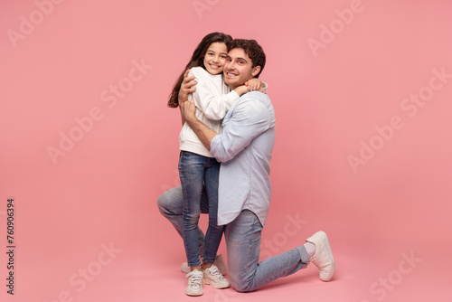 Loving young european father sitting and embracing his preteen daughter and both smiling at camera, posing with child girl on pink studio background, full length