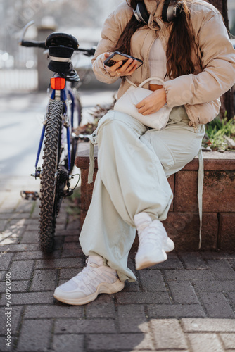 A trendy young adult rests on a city bench with her bicycle, using a smart phone and enjoying music through headphones.