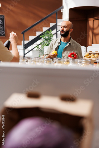 Middle Eastern male customer holds a lemon in one hand and red pepper in another, admiring them both. Young bearded client browsing in an environmentally friendly grocery store.