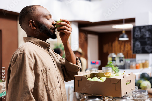 Young man inhales scent of an apple, admiring organic fruits and vegetables. Grocery store supports local producers, offering bulk options and promoting sustainability and zero waste. side view shot.