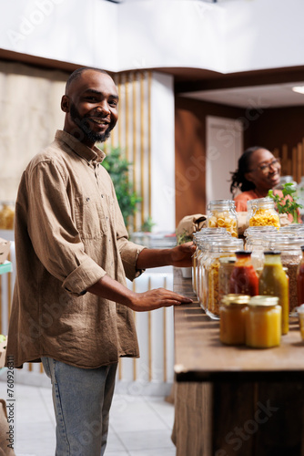 In a modern grocery shop, a happy African American man looks at the camera. The shelves feature reusable packaging, eco friendly products, and a selection of items.