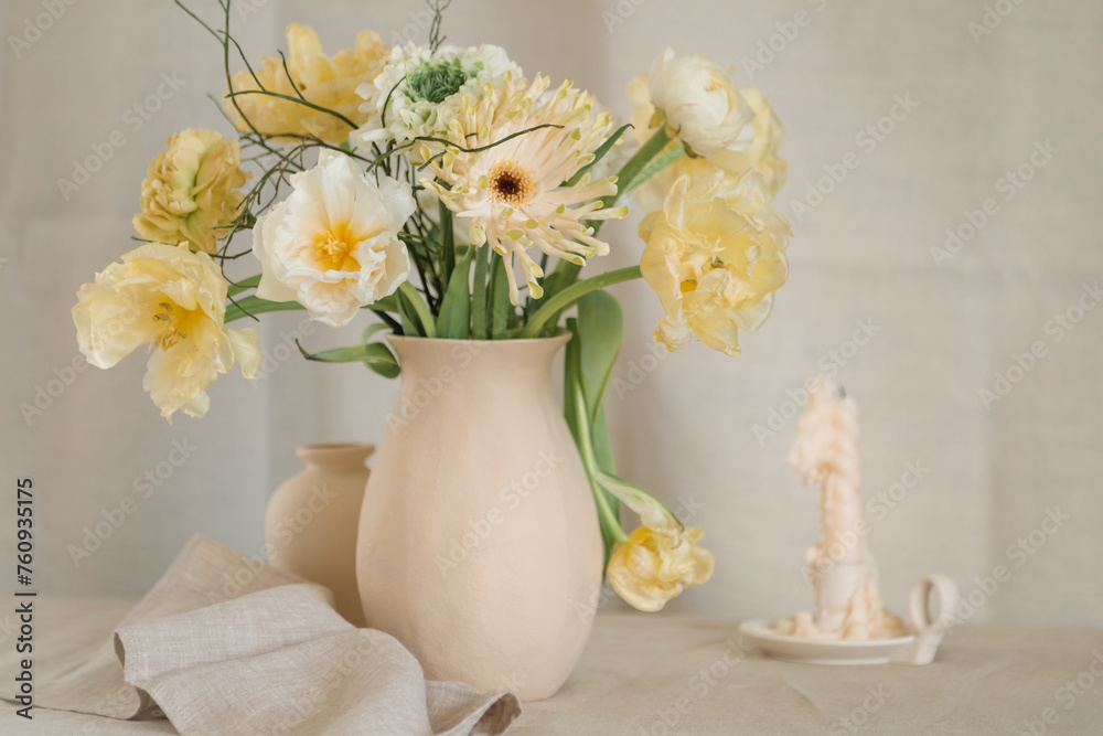Bunch of yellow gerbera, tulip and ranunculus flowers in a vase on a table with a candle and napkin Bunch of yellow gerbera, tulip and ranunculus flowers in a vase on a table with a candle and napkin