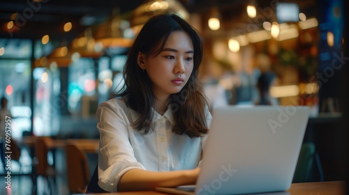 Asian female freelancer deep in concentration, balancing laptop tasks and project notes in a vibrant coffee shop.