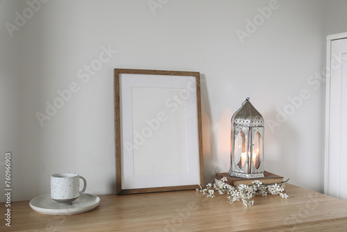 Silver Moroccan lantern, cup of tea, coffee on table. Wooden picture frame, poster mockup. White flowers, blooming prunus tree branches. White wall background, elegant interior. Ramadan Kareem