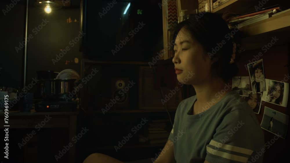 Medium close-up shot of exhausted young Asian woman sitting on bed in micro flat, dimly lit with single light bulb, massaging temples to relieve migraine headache Medium close-up shot of exhausted young Asian woman sitting on bed in micro flat, dimly lit with single light bulb, massaging temples to relieve migraine headache