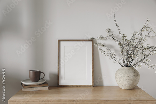Spring interior still life. Cup of coffee, tea on pile of books. White blossoming cherry plum tree branches in ceramic vase on bamboo table. Vertical wooden picture frame, poster mockup. Home decor.