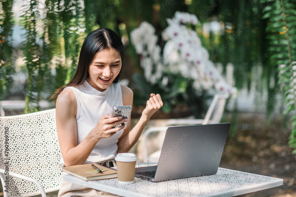 A young woman reacts with delight to her smartphone while taking a break from her laptop work at a garden table, a coffee cup within easy reach. A young woman reacts with delight to her smartphone while taking a break from her laptop work at a garden table, a coffee cup within easy reach.