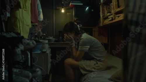 Medium full footage of sad young Chinese woman sitting on bunk bed in cramped dark shoebox apartment, clutching face in hands and crying in despair at hopeless poverty photo