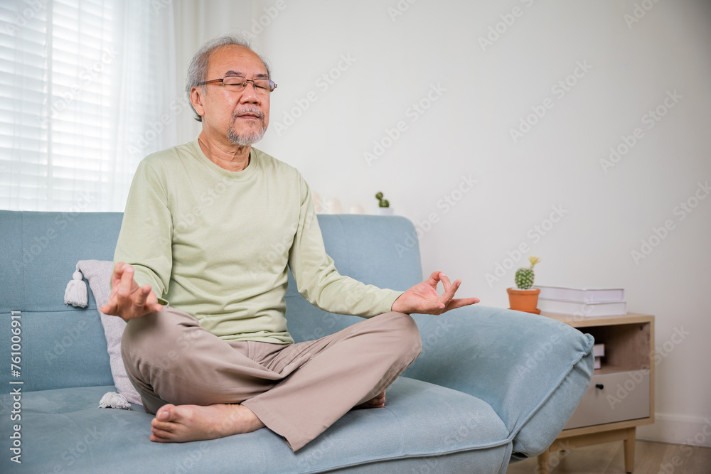 Fototapeta premium Lifestyle senior man sitting on sofa in living room holding hands in mudra practicing home yoga in lotus pose, Elderly relaxed man do physical exercises following healthy on stress free weekend