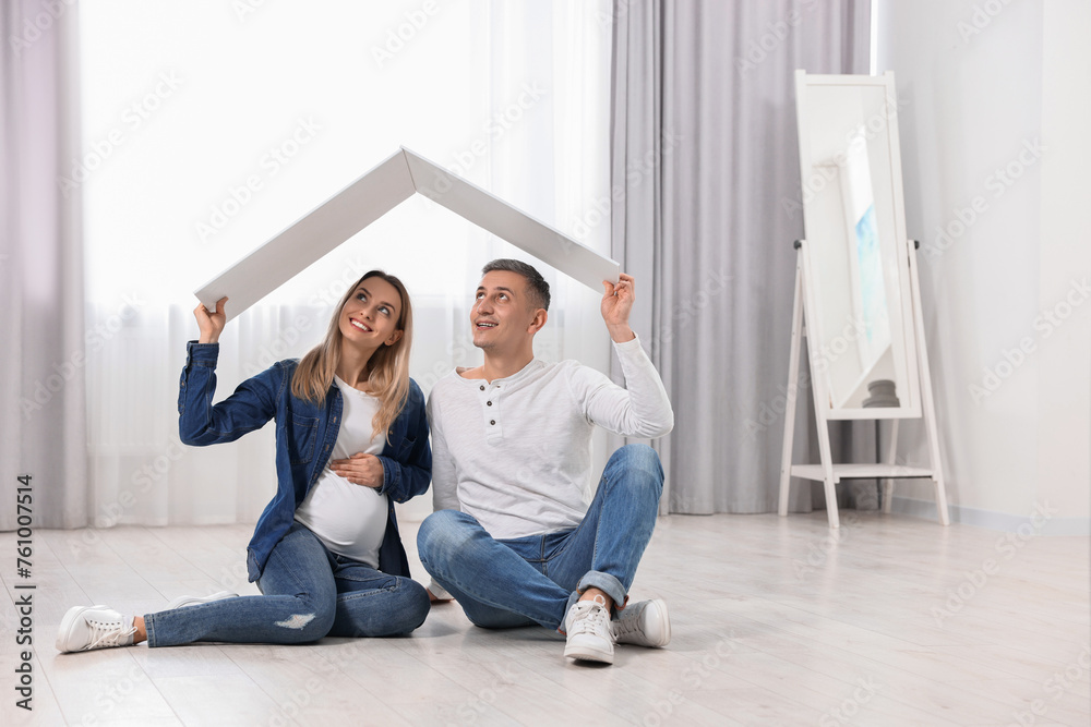 Young family housing concept. Pregnant woman with her husband sitting under cardboard roof on floor indoors. Space for text