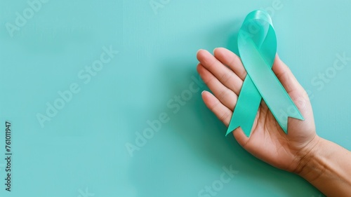 A hand gently holds a teal ribbon against a soft turquoise background symbolizing ovarian cancer awareness photo