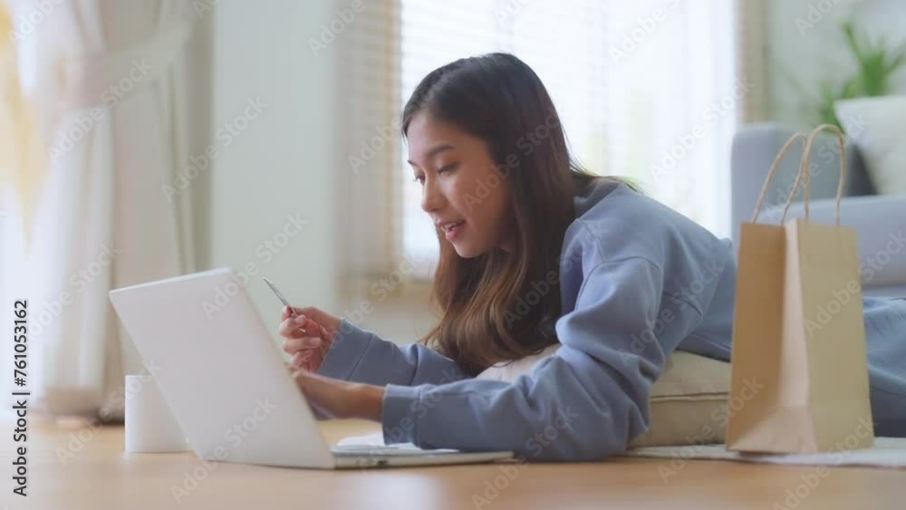 Beautiful young Asian woman working on laptop computer while lying on floor in living room, showing credit card, doing online shopping at home Beautiful young Asian woman working on laptop computer while lying on floor in living room, showing credit card, doing online shopping at home