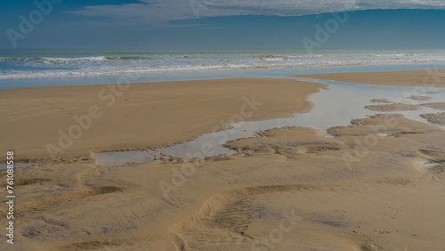 The low tide in the ocean. Puddles and rivulets of water are visible on the sand. The waves of the turquoise ocean roll towards the shore, foaming. Clouds in the blue sky. Madagascar. Morondava. 