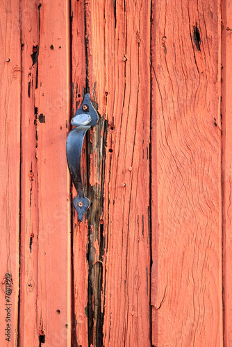 S-grip metal handle and thumb paddle screw mounted on a rundown heavily weathered red wooden door