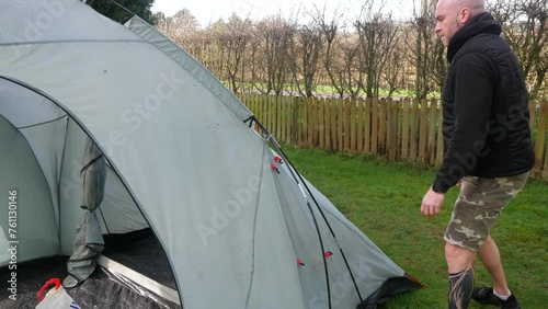A bald man in shorts detaching tent poles from hooks after camping at a campsite whist packing away the tent