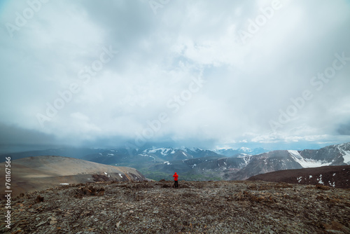 Man in red jacket on high stony pass with view to valley against large snow-capped mountain range in rainy low clouds. Guy among sharp stones on hill. Misty snowy mountains in rain under cloudy sky.