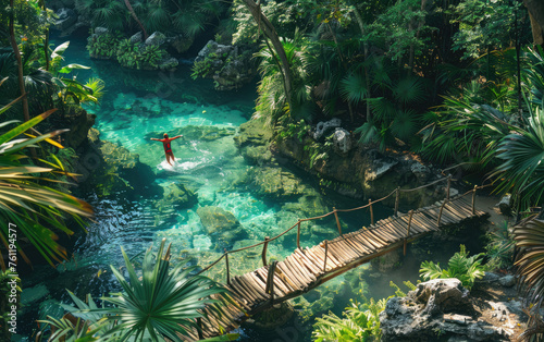 A man jumps into the water from a wooden bridge in a red and black wetsuit. Crystal clear turquoise waters of a Yucatan jungle pool surrounded by lush greenery