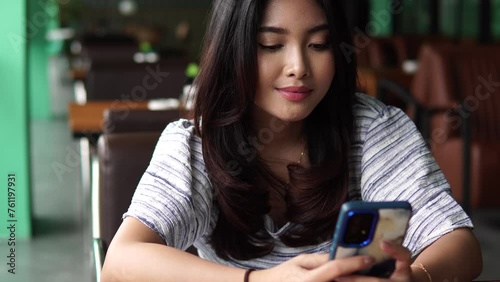Smiling Asian woman using a phone, texting, browsing on social media and internet while sitting down at a restaurant waiting for breakfast