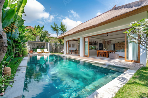 A wide-angle photo of a modern bungalow villa with a pool in a tropical country. Large glass windows and doors overlook the garden and swimming pool, with outdoor seating © Kien