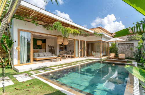 A wide-angle photo of a modern bungalow villa with a pool in a tropical country. Large glass windows and doors overlook the garden and swimming pool, with outdoor seating