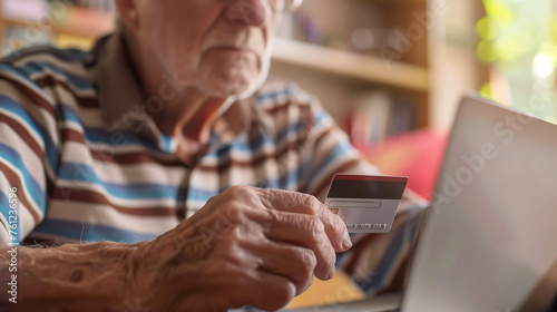 An elderly man, unknowingly falling victim to a scam, enters his banking details online while holding his credit card, unaware of the fraudulent deception targeting him photo