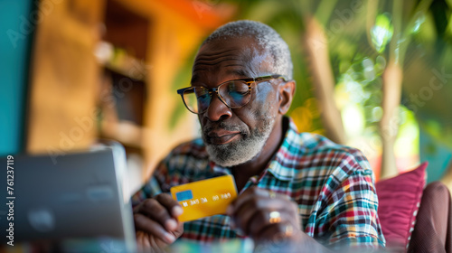 An elderly man, unknowingly falling victim to a scam, enters his banking details online while holding his credit card, unaware of the fraudulent deception targeting him photo