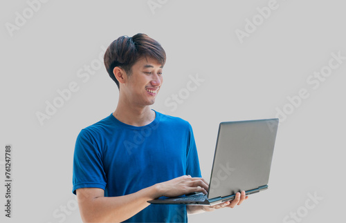 A young Asian man in his 20s wearing a blue t-shirt using a laptop for online remote work isolated on a gray background. Man watching webinars on a laptop, e-learning, e-banking.