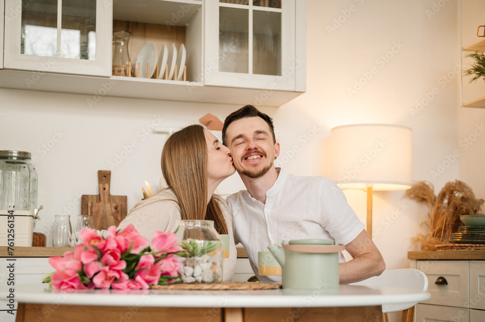Romantic female kisses male, spending time together. Loving young couple having conversation and drink tea in morning. Couple drinking coffee in the kitchen.