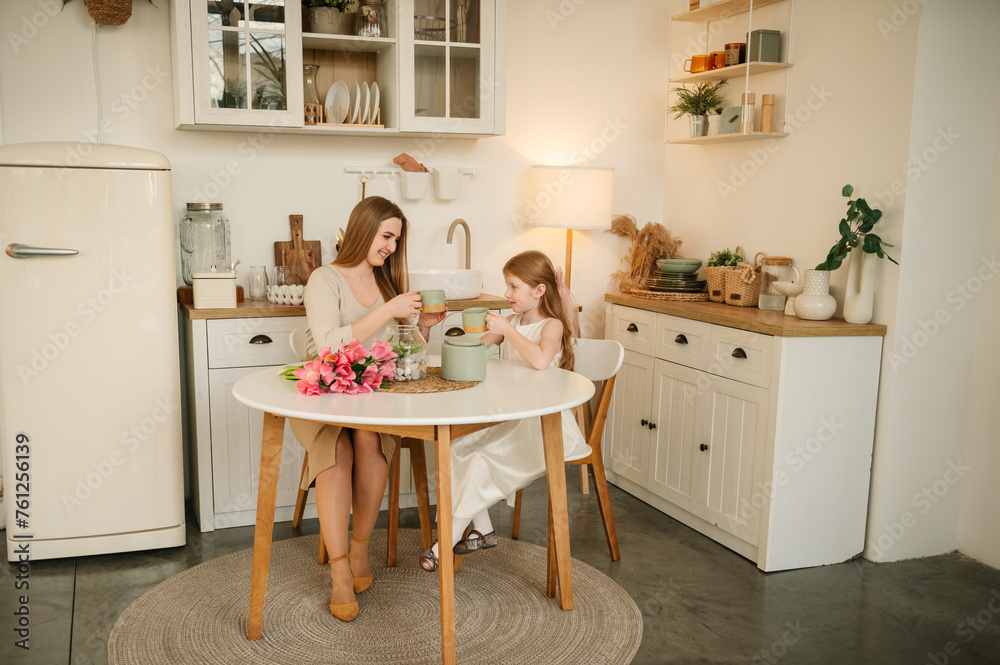 Family communication. Happy mother with daughter drink tea or cocoa sitting in kitchen and having fun. Mom and children spend time together. Child congratulates mother with flowers on Mother's Day. Family communication. Happy mother with daughter drink tea or cocoa sitting in kitchen and having fun. Mom and children spend time together. Child congratulates mother with flowers on Mother's Day.