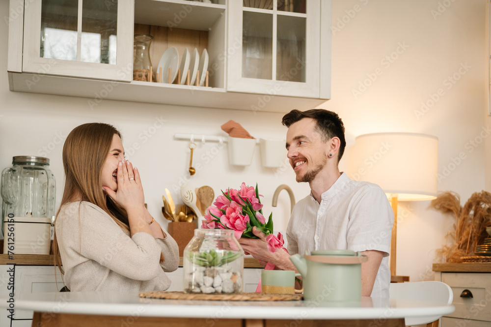 Man congratulate woman and give bouquet of flowers on Women's day. Romantic female and male, spending time together. Loving young couple having conversation and drinking coffee in morning in kitchen.