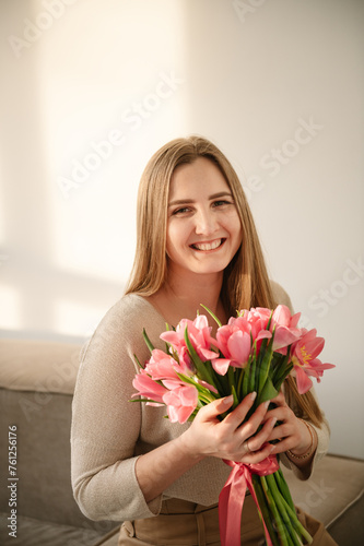 Portrait of a young girl in dress holding hands bouquet of pink tulips. Beautiful woman is smelling a big bunch of spring flowers and sitting on sofa at home. International Women s Day. Looking camera