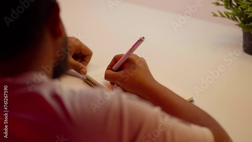 A businessman sits at his home office desk, hands busy writing in a notebook, organizing schedules, tasks, and plans. A close-up highlights the focused male employee jotting down notes with a pen photo