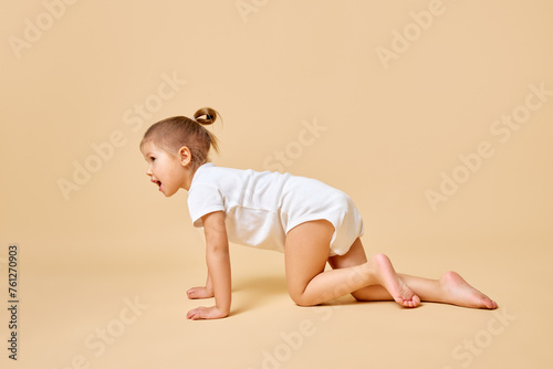 Side view portrait of pretty little baby, girl crawling across floor in cute white romper against beige color background. Concept of childhood, motherhood, life, birth. Copy space for ad