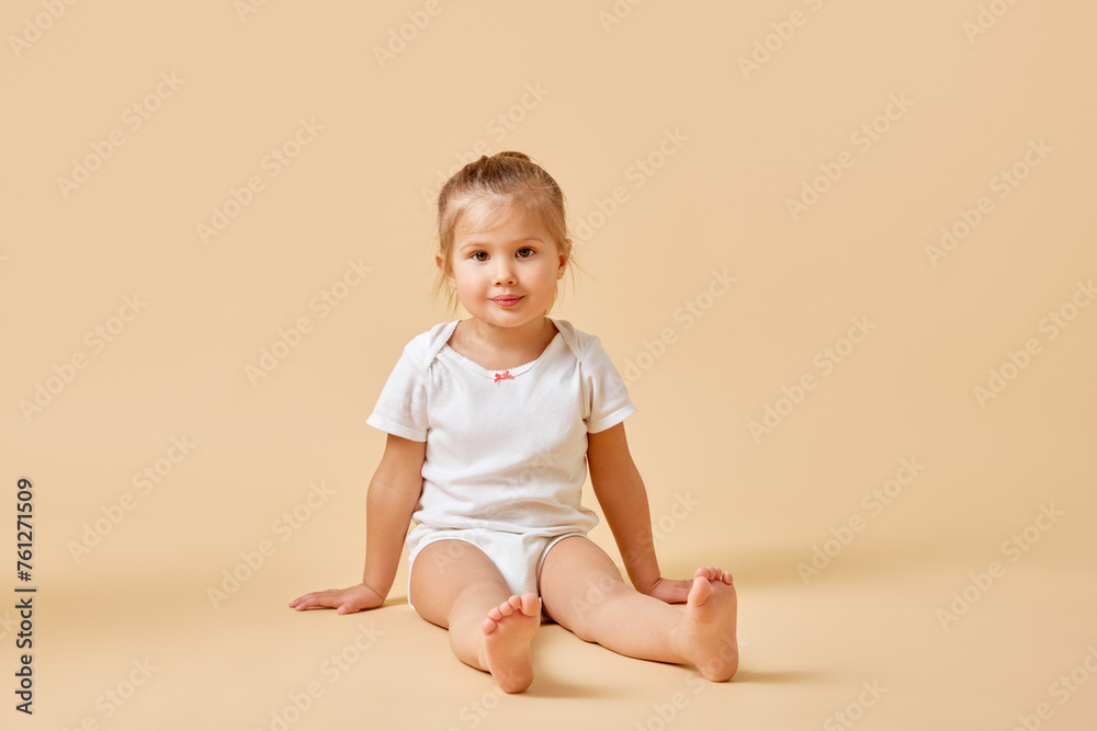 Portrait of joyful little baby in white romper sitting on floor with cute cheerful smile against beige studio background. Concept of childhood, motherhood, life, birth. Copy space for ad Portrait of joyful little baby in white romper sitting on floor with cute cheerful smile against beige studio background. Concept of childhood, motherhood, life, birth. Copy space for ad