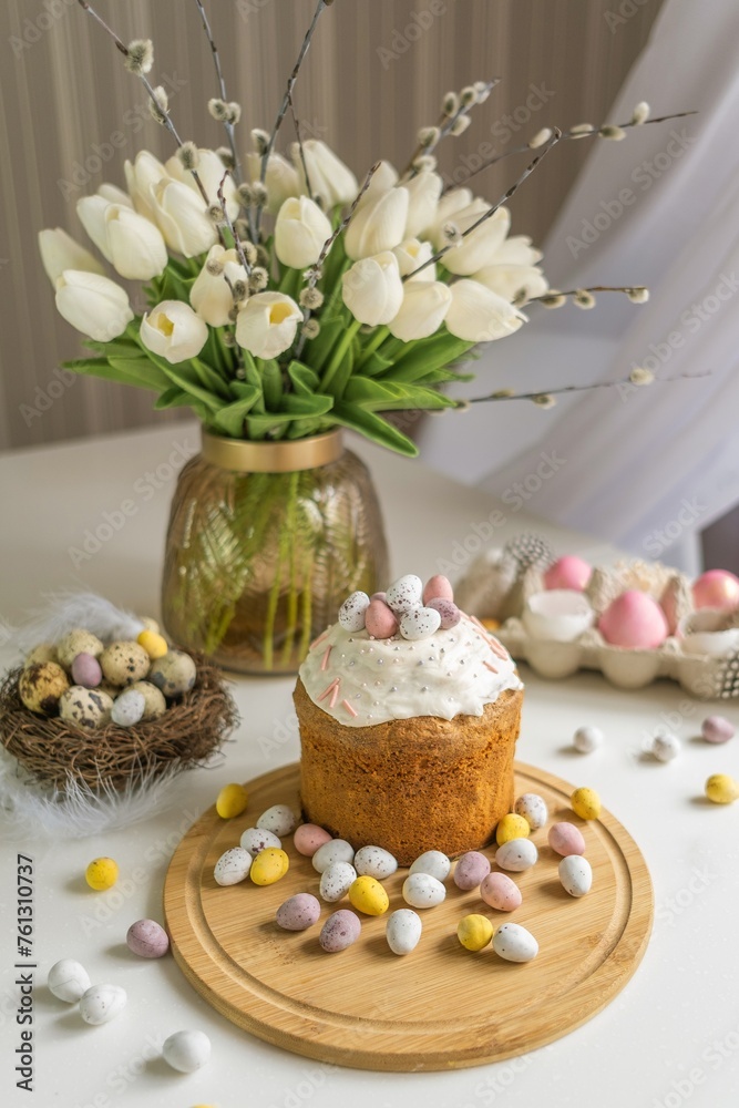 Easter composition on the dining table. Easter cake decorated with colored pastel eggs on a wooden tray. Spring flowers, tulips and willow branches, Easter decor, composition, painted eggs. Soft focus