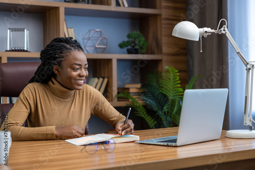 Happy smiling woman sitting at workplace with laptop writing in her organizer photo