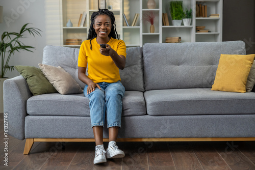 Cute dark-skinned young woman watching tv and holding control panel in hands