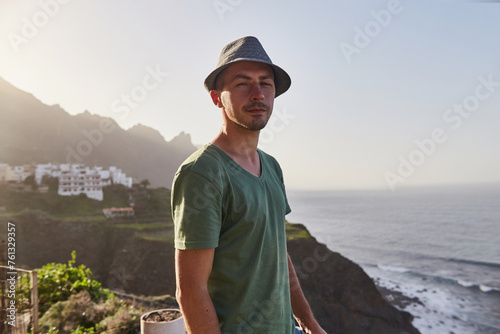 Relaxed male traveler in hat enjoys the coastal view with mountains in the distance