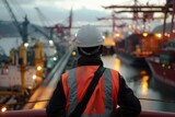 A man with hard hat and safety vest looking cargo ships at the port.