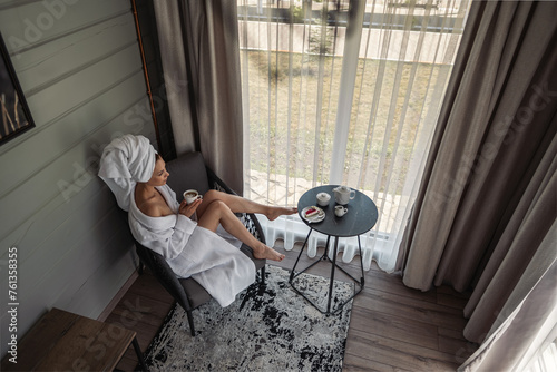 A young girl sits in her robe by the window drinking coffee. A woman in hotel in white robe, resting. Beautiful girl with a towel on her head and robe, in a hotel in an armchair relaxing on vacation.