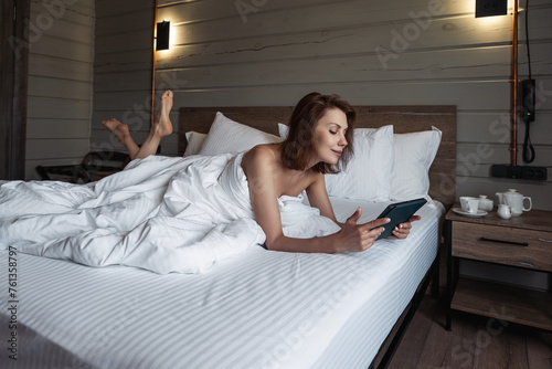 Girl lying in bed using digital tablet In bedroom. Woman lying in bed in hotel room using mobile device for work. Young girl on vacation hotel room on white bed. Person is working while in bed