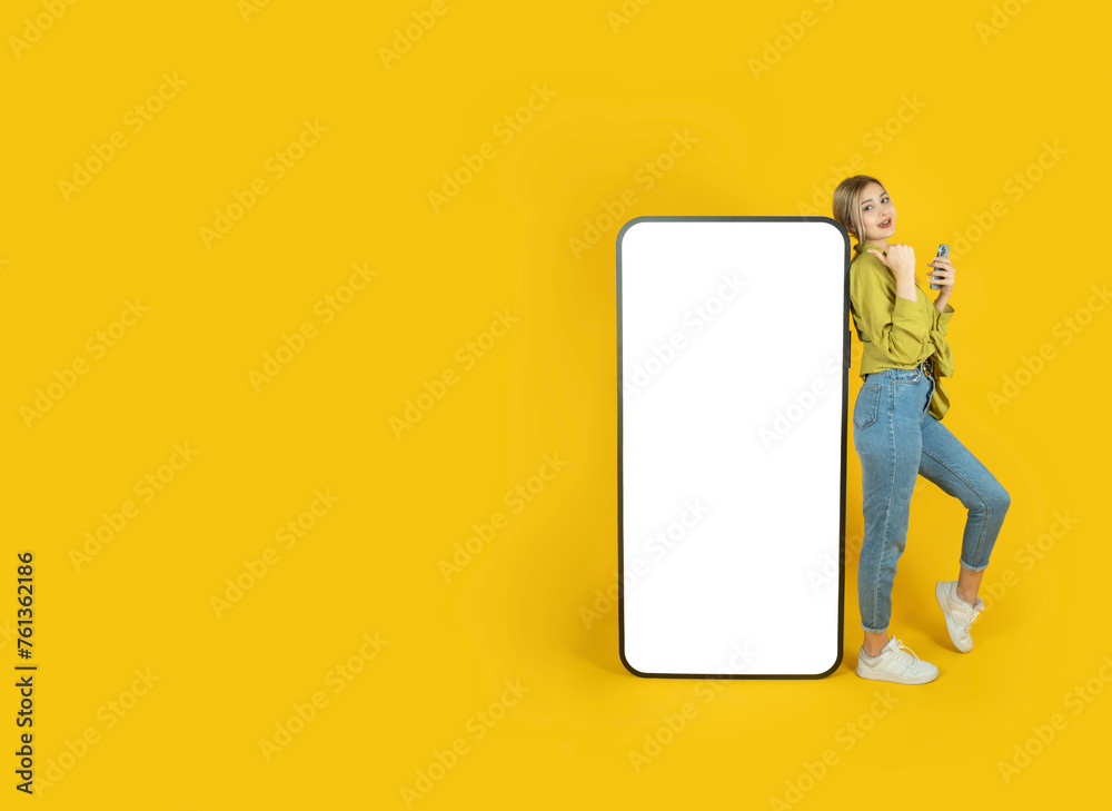 Digital mobile application advertisement concept idea image. Full body young woman 20s leaning big empty screen cell phone direct thumb display touchscreen mock up. Yellow studio background copy space Digital mobile application advertisement concept idea image. Full body young woman 20s leaning big empty screen cell phone direct thumb display touchscreen mock up. Yellow studio background copy space