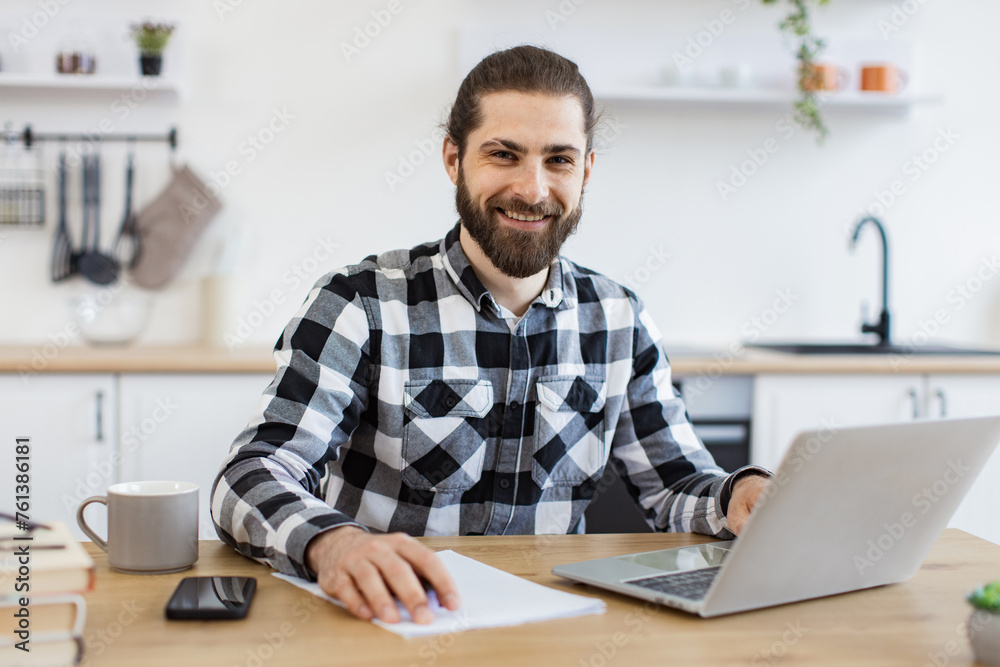 Efficient business owner improving contract while checking data on portable computer.Handsome Caucasian man wearing checkered shirt writing on document while staying at home during midday. Efficient business owner improving contract while checking data on portable computer.Handsome Caucasian man wearing checkered shirt writing on document while staying at home during midday.